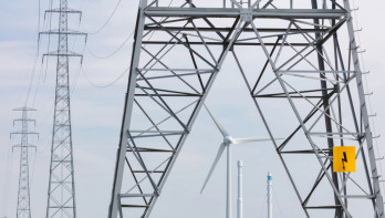 Groep Nederlandse hoogspanningsmasten met een windturbine in het landschap.