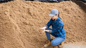 Vrouw die gehurkt zit naast een grote hoop bierbostel in een industriële of agrarische omgeving.