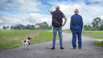 Twee Nederlandse boeren aan de achterkant kijken uit over een landweg en weilanden aan de horizon.