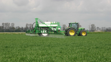 Een groene tractor met veldspuit brengt gewasbeschermingsmiddel aan op een dicht, groen graanperceel onder een bewolkte lucht.