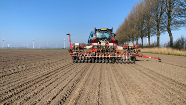 Un tracteur équipé d'un semoir sème un champ à la campagne, avec des éoliennes en arrière-plan.