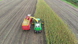 Aerial view of a forage harvester harvesting maize and loading the chopped crop into a multi-wheeled trailer.