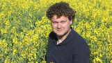 Man met krullend haar in een donkerblauwe polo staat in een uitgestrekt veld met bloeiende gele koolzaadbloemen op een zonnige dag.