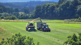 Een hakselaar vult een aanhanger achter een tractor met vers gemaaid gras op een groen weiland, omringd door bossen en heuvels.